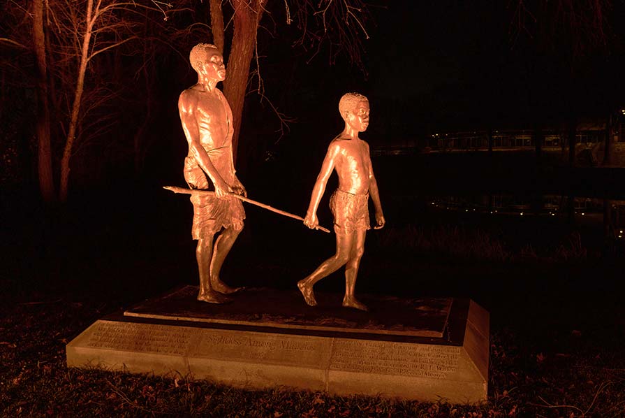 Photo of a bronze statue where a young boy uses a walking stick to guide a blind man, illuminated with orange light.