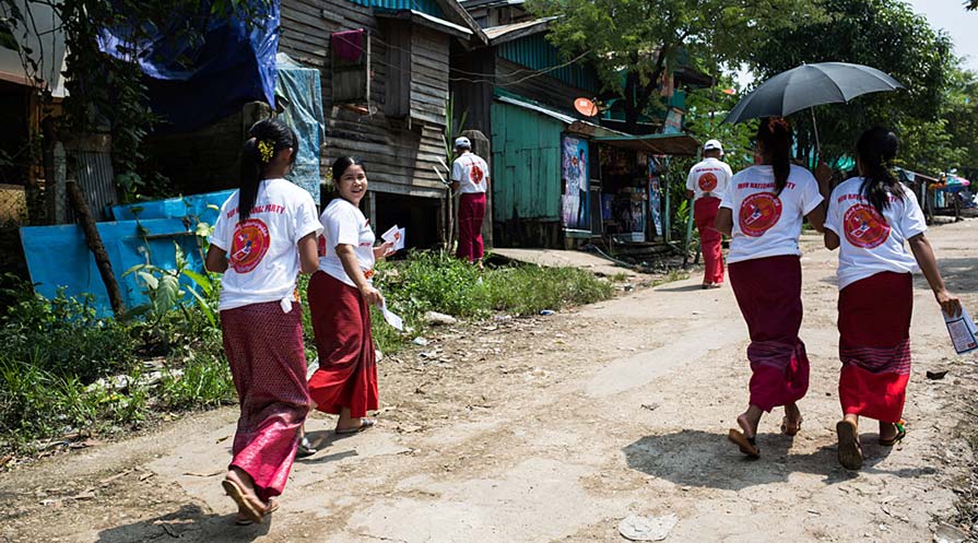 Photo of a group of women in matching election t-shirts walk a dirt road distributing flyers and pamphlets.