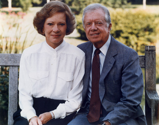 Portrait of Rosalynn and Jimmy Carter sitting outside on a wooden bench.