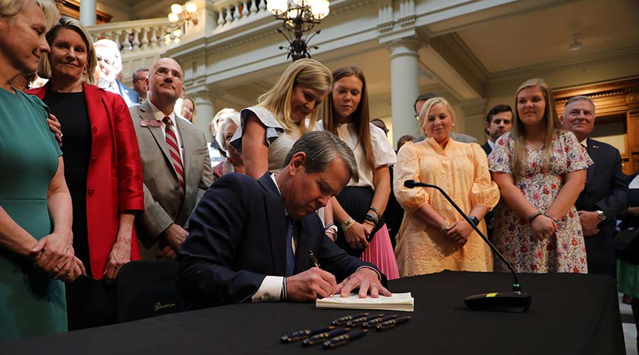Brian Kemp sits at a table while onlookers stand behind him as he signs the parity act.