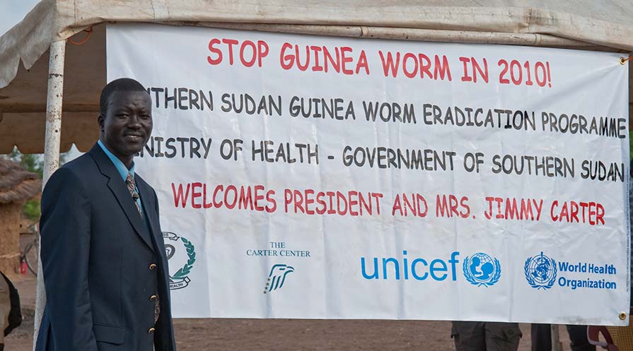 Makoy Samuel Yibi parado frente a un cartel de Stop Guinea Worm.