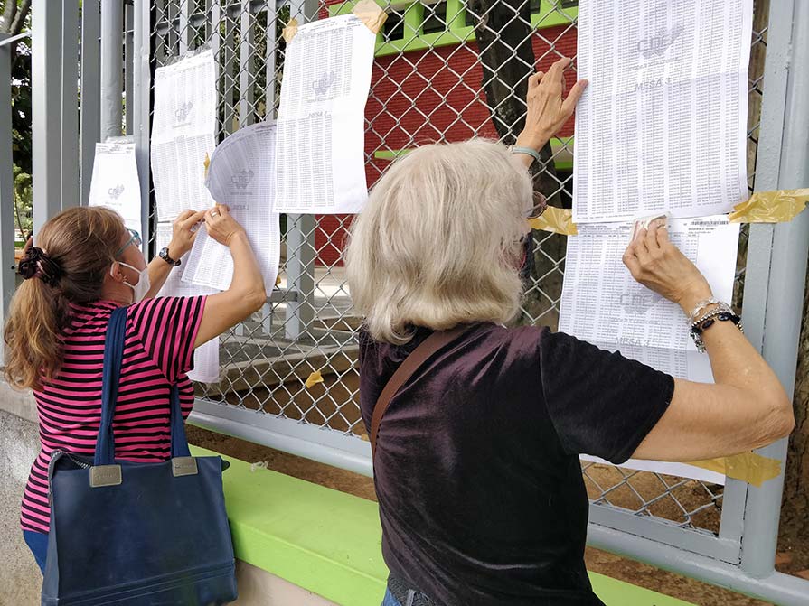 Two women check a paper list that hangs on a fenced section of a wall.