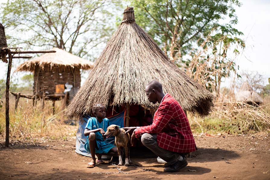 A man, child, and small dog kneel in front of a village hut.