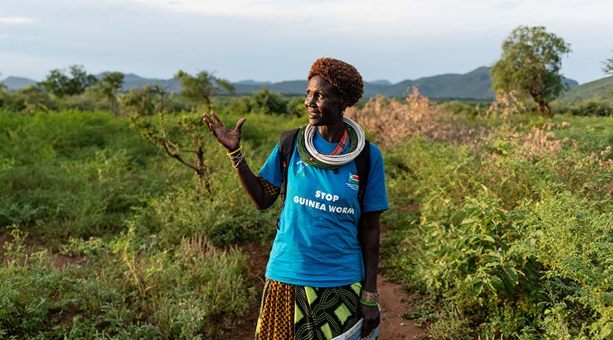 Medium shot of Regina Natube standing in grassy terrain wearing a blue shirt that reads Stop Guinea Worm.