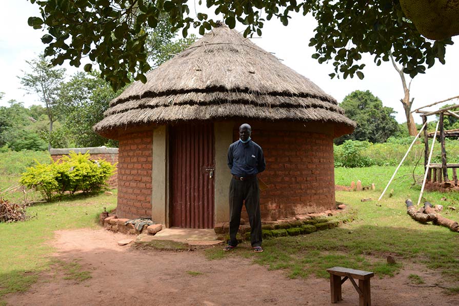 Fred stands solo in front of a thatched roof structure in a village.